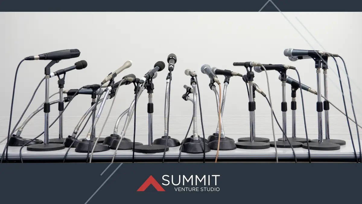 A lineup of assorted microphones on stands arranged in a semicircle on a table, suggesting a press conference setup. The Summit Venture Studio logo is displayed at the bottom of the image.