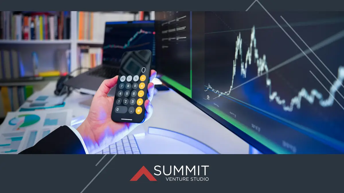 Person holding a smartphone calculator in front of computer monitors displaying financial graphs and data, with printed charts on the desk.