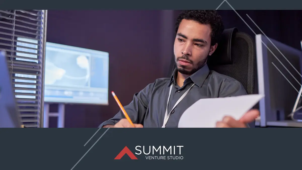 Entrepreneur analyzing research data and documents at a computer workstation in a modern office environment with Summit Venture Studio branding.